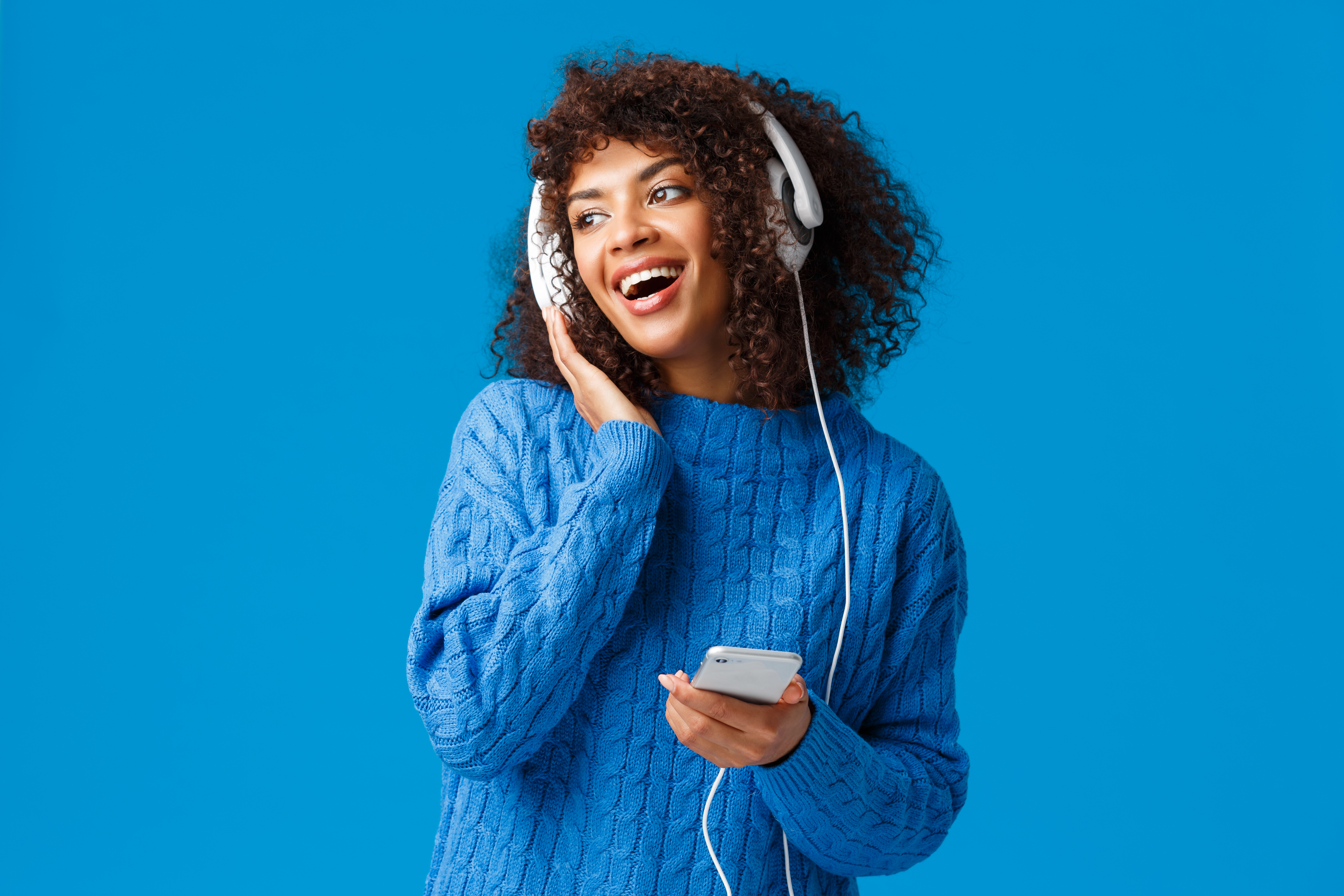 charismatic modern young attractive african american girl with afro haircut, listening music in headphones looking out window snowy weather, holding smartphone, smiling and singing